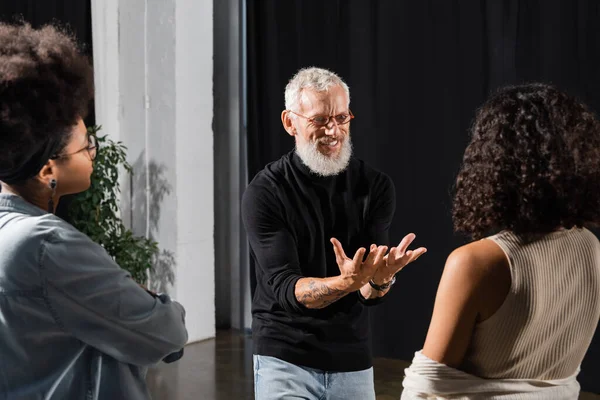 Smiling grey haired art director in eyeglasses talking and gesturing near interracial actresses in theater school — Photo de stock