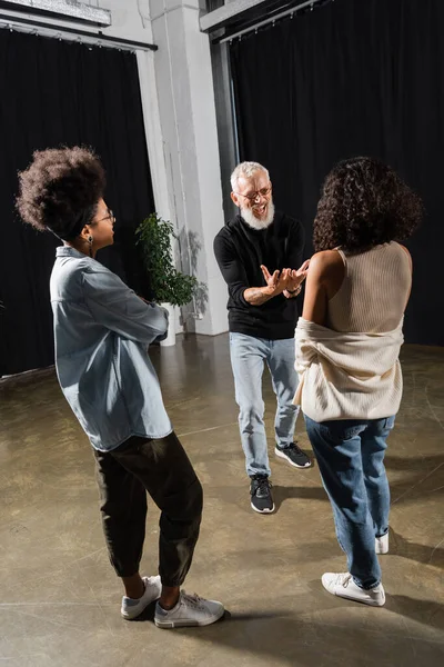 Excited bearded producer gesturing and talking near multiethnic actresses in acting school - foto de stock