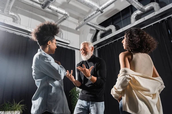 Positive and emotional acting skills teacher gesturing near young interracial women in theater. Translation of tattoo: om, shanti, peace — Stock Photo
