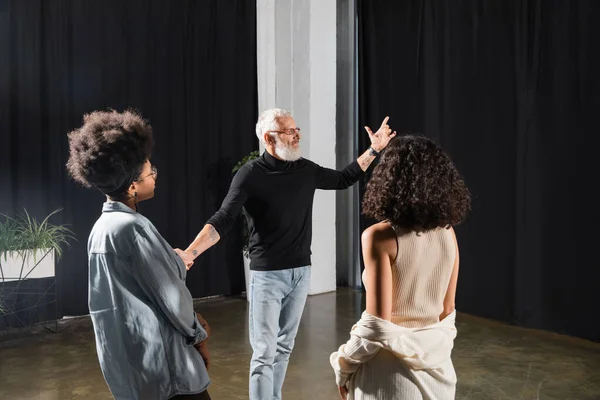 Bearded middle aged art director pointing with hand near interracial actresses during lesson in theater. Translation of tattoo: kanji, danger - foto de stock