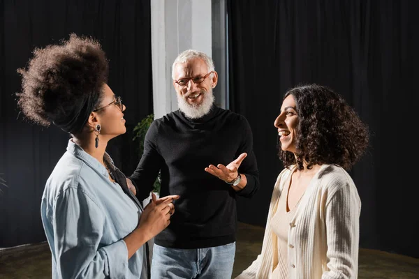 Positive grey haired producer pointing with hand while talking to cheerful interracial actresses in theater — Stock Photo