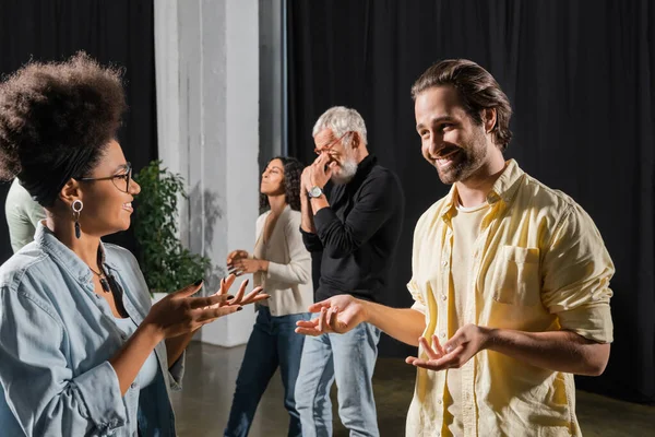 Smiling brunette man talking to african american woman near interracial colleagues in acting school — Foto stock