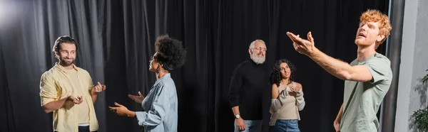 Young redhead man posing with outstretched hand near interracial actors talking in acting school, banner — стоковое фото
