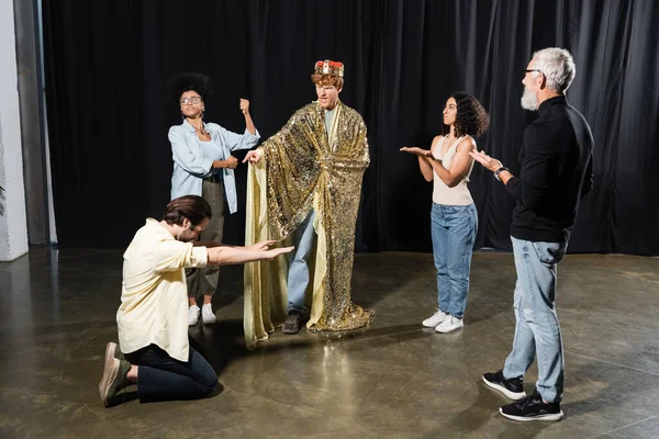Redhead man in king costume rehearsing role near multiethnic actors and art director in acting school - foto de stock