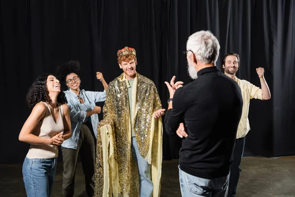 Cheerful redhead man in king costume near multiethnic actors and art director on stage in theater — Foto stock
