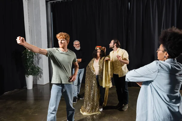 Multiracial woman in queen costume and redhead man with outstretched fist rehearsing scene in acting skills school - foto de stock
