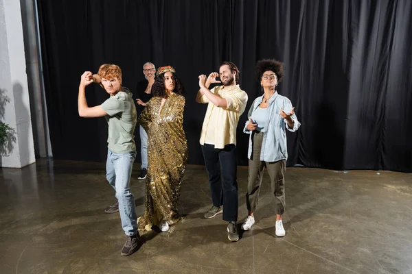 Multiracial woman in queen costume rehearsing with young multiethnic actors near art director smiling on blurred background — Stockfoto