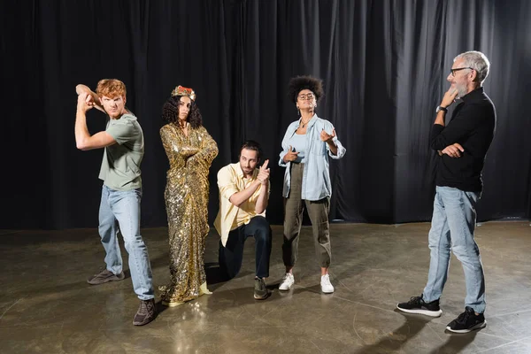 Grey haired art director looking at multiracial woman in queen costume standing with crossed arms near actors rehearsing guards roles — Stock Photo