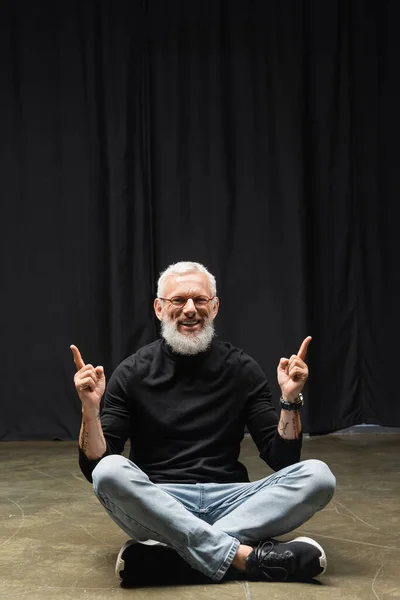 Cheerful tattooed actor pointing up with fingers while sitting on theater stage with crossed legs. Translation of tattoo: om, shanti, peace - foto de stock