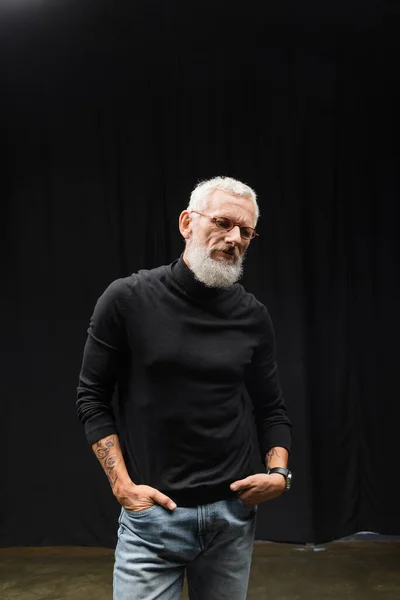 Mature tattooed actor in eyeglasses and black turtleneck standing with hands in pockets of jeans in theater - foto de stock