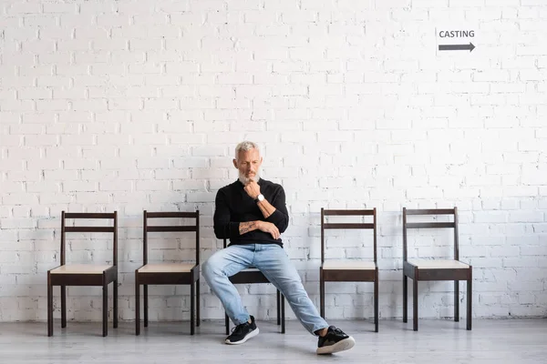 Full length of pensive bearded man sitting in hall near white wall and waiting for casting. Translation of tattoo: om, shanti, peace — Stock Photo