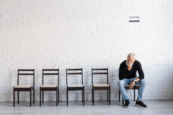 Full length of pensive man in black turtleneck and jeans waiting for casting on chair near white wall — Stock Photo