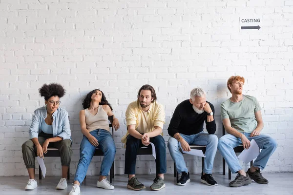 Bored multicultural actors holding screenplays while sitting on chairs and waiting for casting — Stock Photo