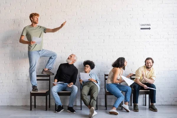 Redhead man standing on chair and rehearsing near multiethnic actors sitting and waiting for casting — Stockfoto