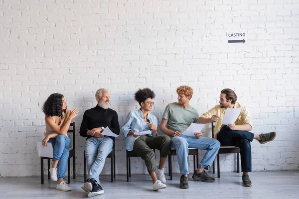 Positive multicultural actors holding screenplays and talking while sitting on chairs and waiting for casting — Stockfoto