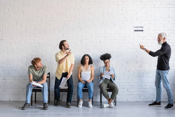 Bearded art director calling brunette man near multiethnic actors sitting on chairs and waiting for casting — Foto stock