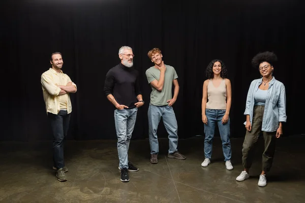 Smiling bearded art director standing with hands in pockets near young multiethnic actors on stage in theater — Stock Photo
