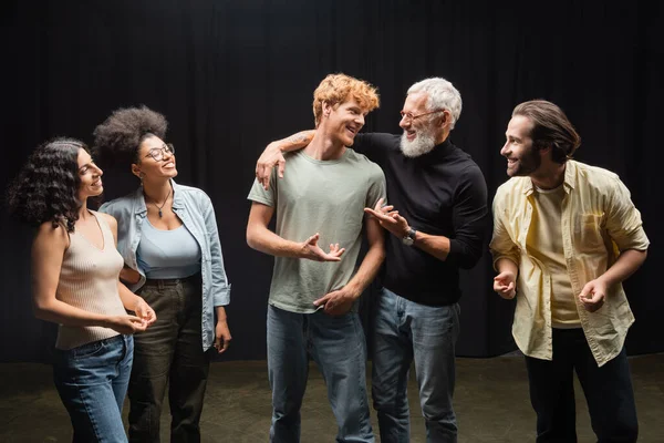 Happy producer talking to redhead man near smiling interracial actors on theater stage — Foto stock