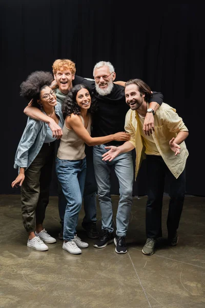 Young and joyful interracial actors looking at camera while embracing bearded producer in theater — Foto stock