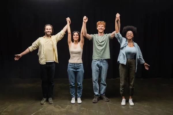 Full length of young and joyful interracial actors holding raised hands and smiling at camera in theater — стоковое фото