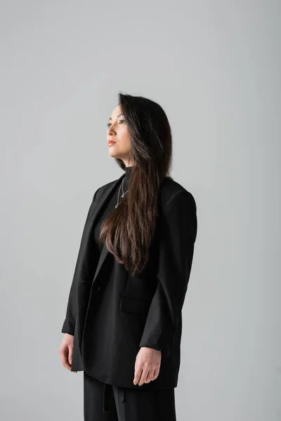 Asian brunette woman in black blazer looking away isolated on grey — стоковое фото