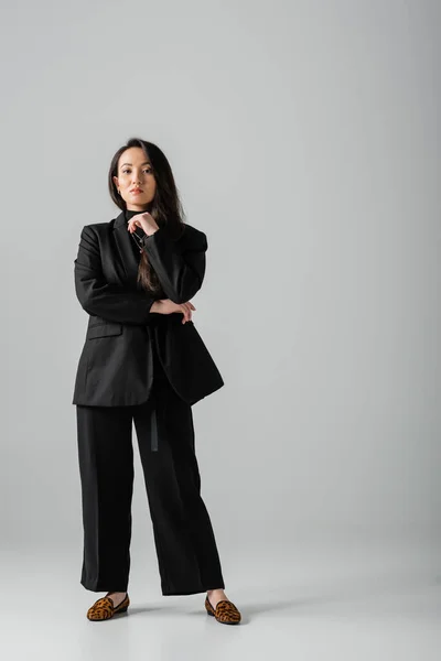 Full length of brunette and asian businesswoman in black suit standing on grey — стоковое фото