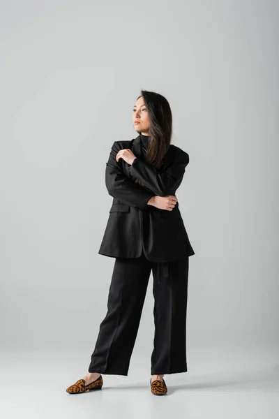 Full length of young asian businesswoman in black suit standing and looking away on grey — стоковое фото