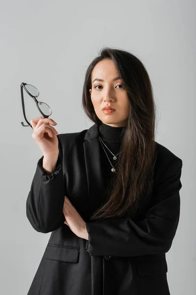 Portrait of young asian businesswoman in black suit holding glasses and looking at camera isolated on grey — Stock Photo