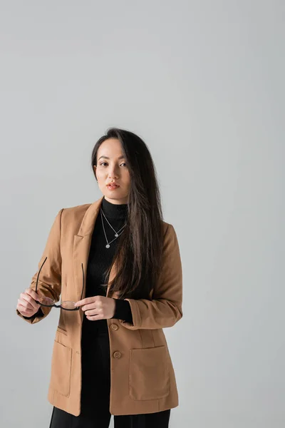 Young asian businesswoman in beige blazer holding glasses and looking at camera isolated on grey — Stockfoto