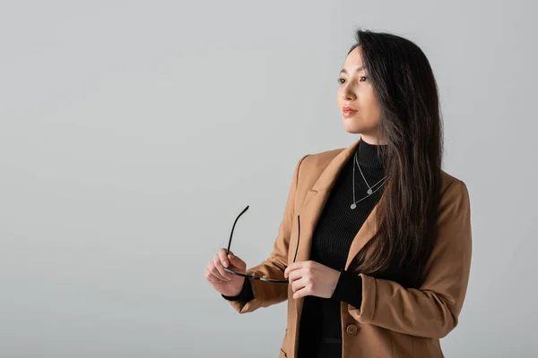 Asian businesswoman in beige blazer holding glasses and looking away isolated on grey — Fotografia de Stock
