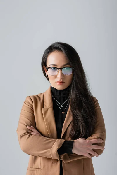 Portrait of brunette asian businesswoman in beige blazer and glasses standing with crossed arms isolated on grey — Stock Photo