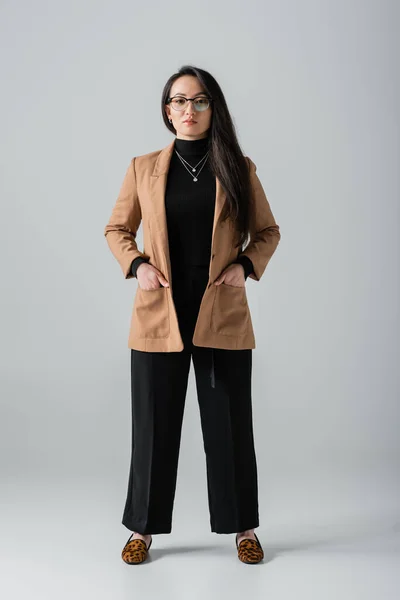 Повна довжина молодої азіатської бізнес-леді в бежевих блейзер і чорних штанях, що стоять руками в кишенях на сірому — стокове фото