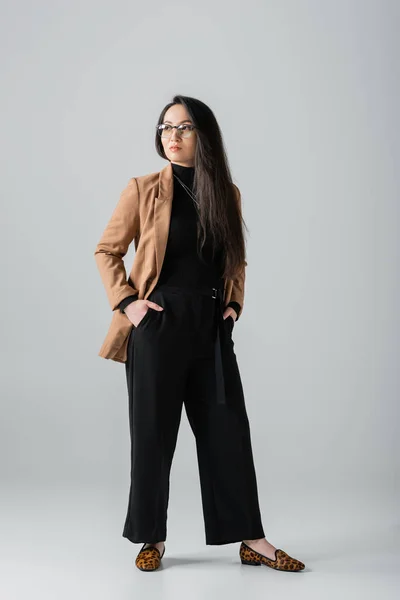 Full length of confident asian businesswoman in beige blazer and black pants standing with hands in pockets on grey — Foto stock