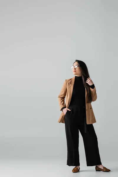 Full length of confident asian businesswoman in beige blazer and black pants standing with hand in pocket on grey — Foto stock