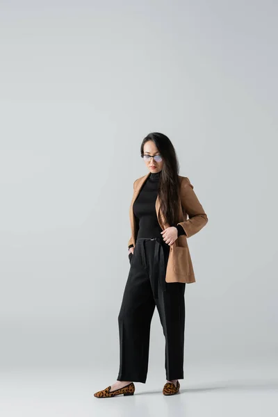 Повна довжина азіатської бізнес-леді в бежевих та чорних штанях, що стоять з рукою в кишені на сірому — стокове фото