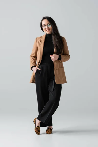 Full length of happy asian businesswoman in beige blazer and black pants standing on grey — стоковое фото