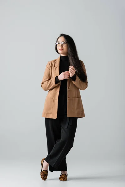 Повна довжина молодої азіатської бізнес-леді в бежевих та чорних штанях, що стоять з схрещеними ногами на сірому — стокове фото