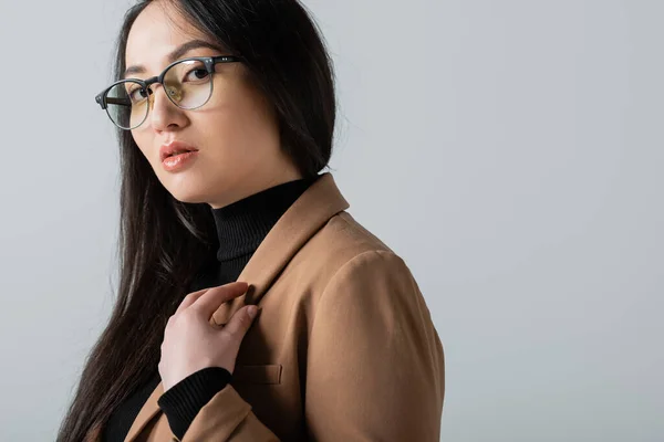 Young asian woman in glasses and blazer looking at camera isolated on grey — Stock Photo