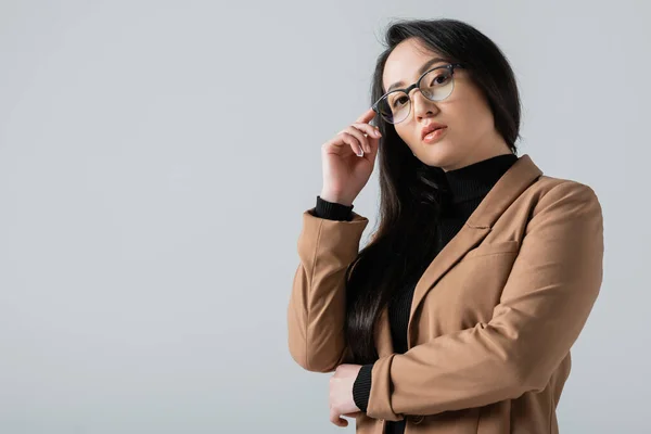 Young asian woman in beige blazer looking at camera while adjusting glasses isolated on grey — Photo de stock