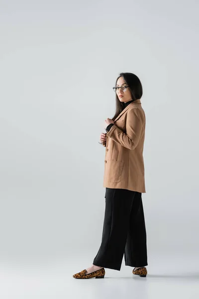 Full length of young asian woman in beige blazer and black pants posing on grey — Stock Photo