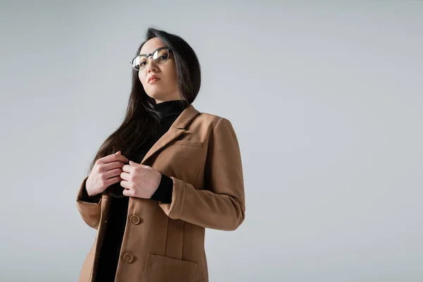 Low angle view of asian businesswoman in beige blazer and black turtleneck looking at camera isolated on grey — Stock Photo