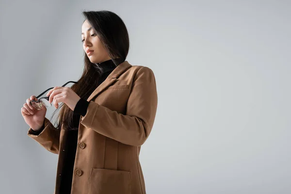 Low angle view of asian businesswoman in beige blazer and black turtleneck looking at glasses isolated on grey — Fotografia de Stock