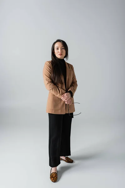 Full length of young asian woman in beige blazer and black pants holding glasses on grey — Fotografia de Stock