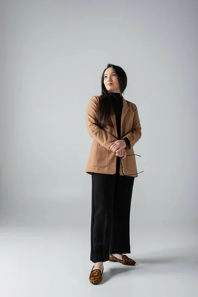 Full length of brunette asian woman in beige blazer and black pants holding glasses on grey — стоковое фото