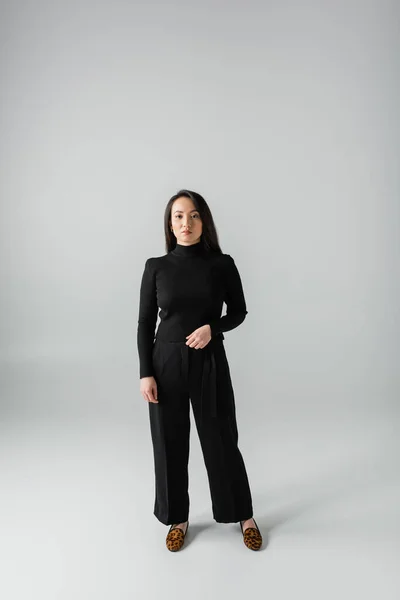 Full length of young asian woman in black pants and turtleneck standing on grey - foto de stock