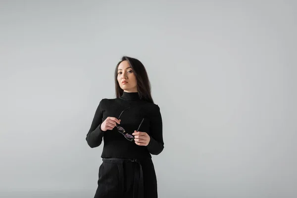 Brunette asian businesswoman in black turtleneck looking at camera while holding glasses isolated on grey — Fotografia de Stock