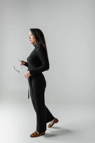 Full length of brunette asian woman in black outfit and loafers with animal print holding glasses on grey — Photo de stock