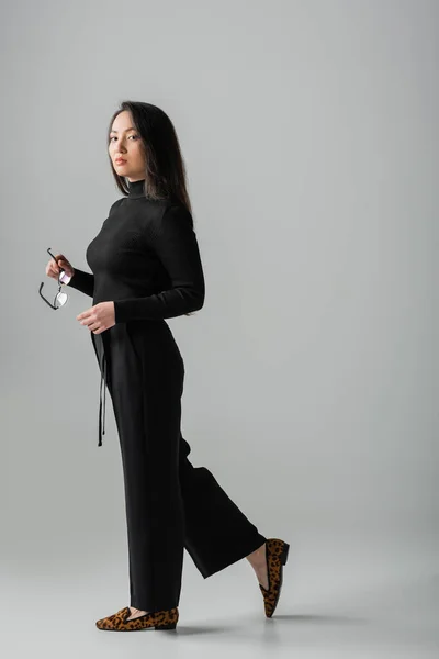 Full length of brunette asian woman in stylish black outfit and loafers standing with glasses on grey — Stock Photo