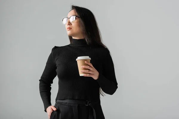 Brunette asian woman in stylish black turtleneck holding paper cup while posing with hand in pocket isolated on grey — стоковое фото