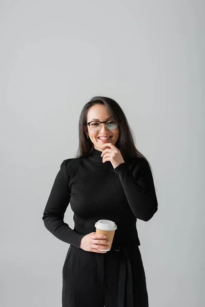 Pleased asian woman in black turtleneck and glasses holding coffee to go isolated on grey — Stock Photo